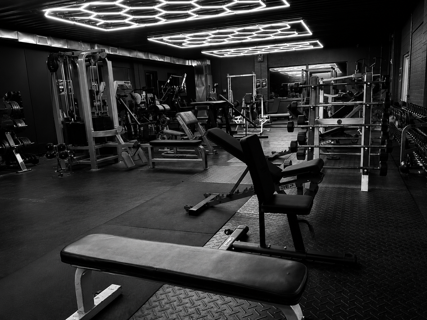 The training floor at The Gym: hexagonal LED ceiling panels above power racks, benches and a full wall of dumbbells